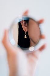 A hand holds a circular mirror reflecting a person's ear wearing a gold earring with a clear stone.